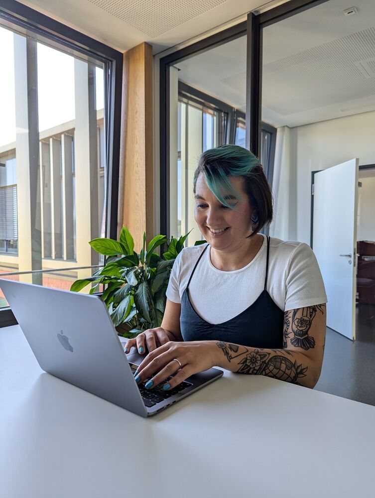 Eine junge Frau mit tätowierten Armen und blauen Haaren arbeitet am Laptop in einer Bibliothek. 