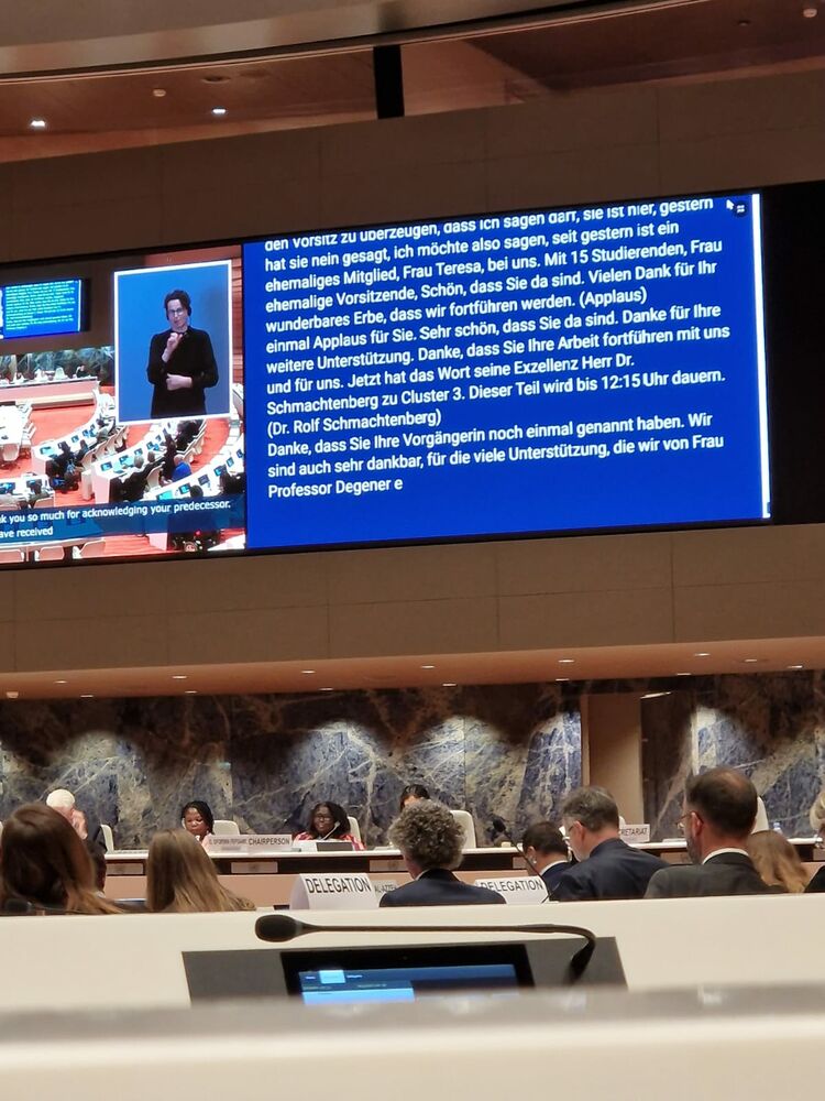 Blick in den Sitzungssaal bei den Vereinten Nationen, mit dem Präsidium und einen großen Screen für Schrift- und Gebärdensprachdolmetschung. Es ist der Ausschnitt mit der Begrüßung zu lesen.